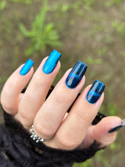 Heart of the Ocean by Glitch Lacquer on five square nails comparing magnetized and unmagnetized finishes, deep blue jelly with blue-to-purple magnetic shift and blue glitters, light-medium skin tone, bright daylight.