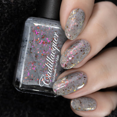 Cadillacquer Daydreaming grey red chameleon iridescent crelly nail polish, bottle held with five-nail swatch, light skin, studio lighting