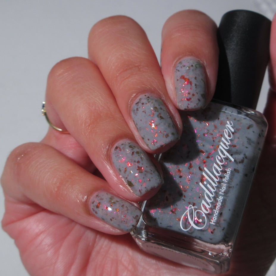 Cadillacquer Daydreaming grey red chameleon iridescent crelly nail polish, bottle held with four-nail swatch, light skin, bright daylight lighting