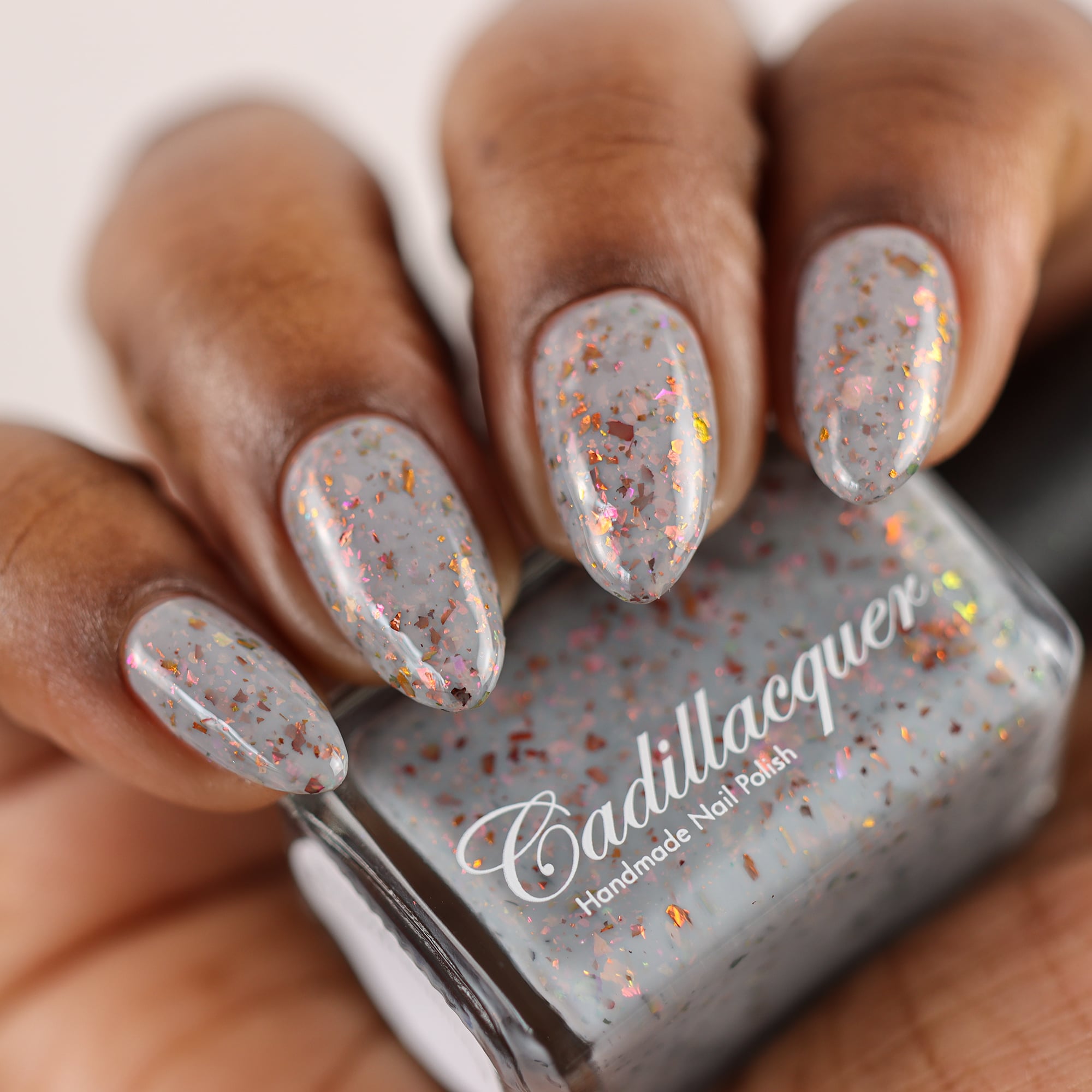 Cadillacquer Daydreaming grey red chameleon iridescent crelly nail polish, bottle held with four-nail swatch, deep skin, studio lighting