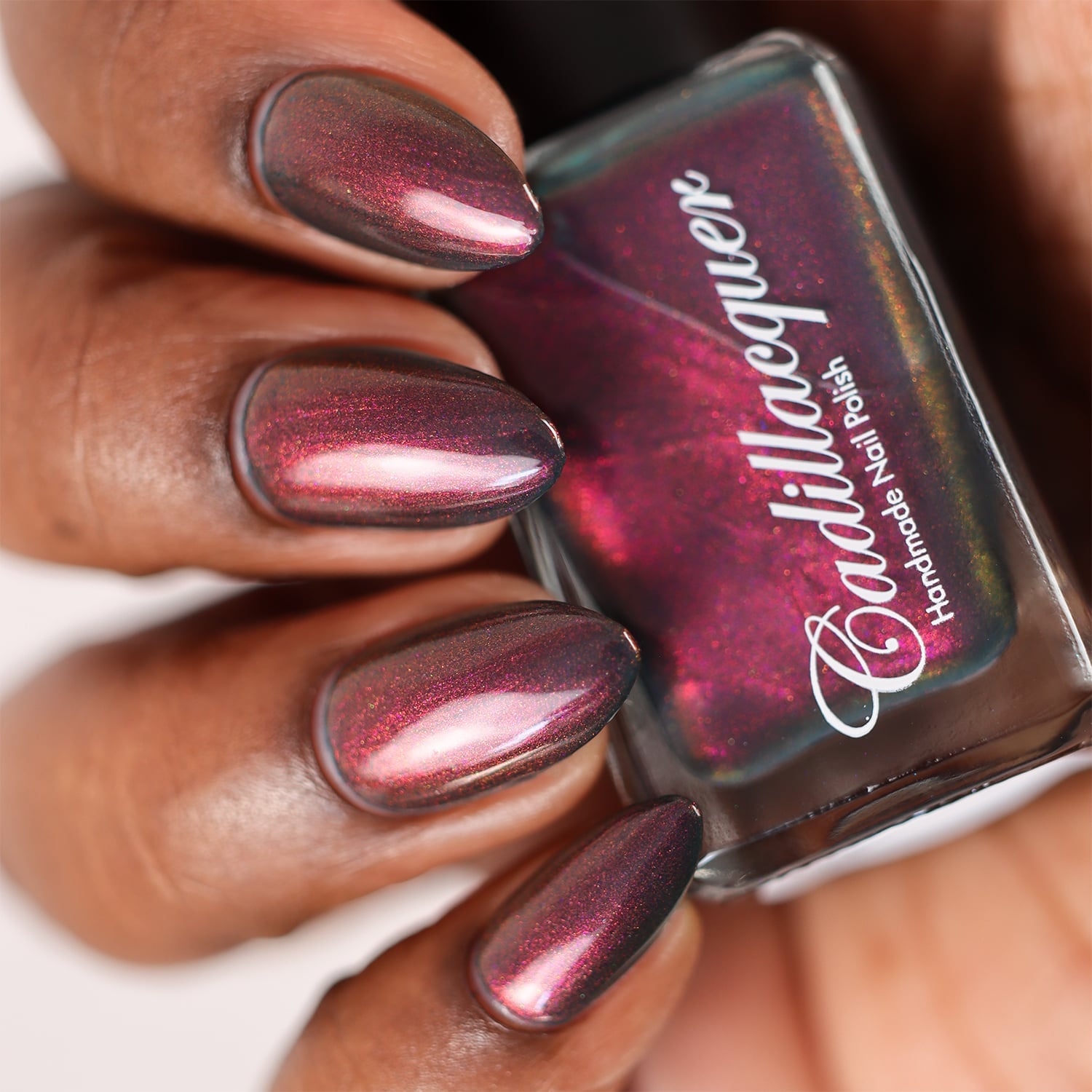 Multichrome shimmer flakie Cadillacquer nail polish in Fear What You Will Become, shown in bottle held in hand with five almond-shaped nails swatched, shifting from deep blackened teal base through magenta-crimson to orange-gold shimmer depending on angle, on deep skin tone, under studio lighting, swatched by @doseoflolade