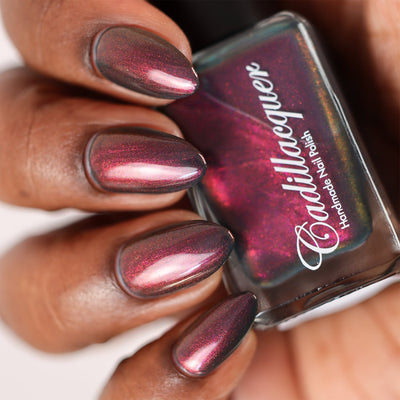 Multichrome shimmer flakie Cadillacquer nail polish in Fear What You Will Become, shown in bottle held in hand with five almond-shaped nails swatched, shifting from deep blackened teal base through magenta-crimson to orange-gold shimmer depending on angle, on deep skin tone, under studio lighting, swatched by @doseoflolade