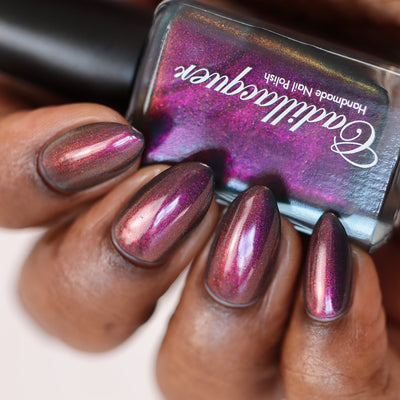 Multichrome shimmer Cadillacquer nail polish in Fear What You Will Become, shown in-bottle held against swatched almond nails on rich skin tone, nails shift from deep dusty plum-brown at rest through vivid magenta and copper-orange at angles, bottle face blazes bright magenta with gold shimmer while bottle edge reveals the murky teal base, studio lighting, swatched by @doseoflolade
