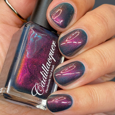 Multichrome shimmer Cadillacquer nail polish in Fear What You Will Become, shown in-bottle held in hand alongside four swatched short oval nails, the bottle face blazes magenta-to-orange while the bottle edge and nail body read murky dark teal, with magenta shimmer flashing at angular highlights on the nails, light-medium skin tone, studio lighting, swatched by @heeymercedes