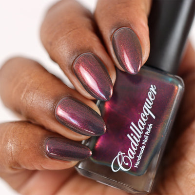Multichrome shimmer Cadillacquer nail polish in Fear What You Will Become, swatched on almond-shaped nails held around a polish bottle, showing a deep dark burgundy base that shifts through magenta to copper-orange shimmer, on rich skin tone, studio lighting, swatched by @doseoflolade