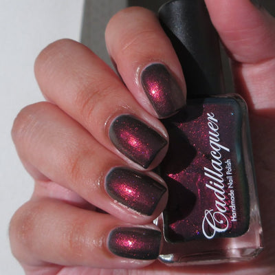 Shimmer Cadillacquer nail polish in Fear What You Will Become, swatched on eight squoval nails held alongside the bottle, showing a very dark blackened-plum base packed with vivid magenta-to-orange-red shimmer that shifts dramatically with angle, on light-medium skin, in bright daylight, swatched by @_always_polished_