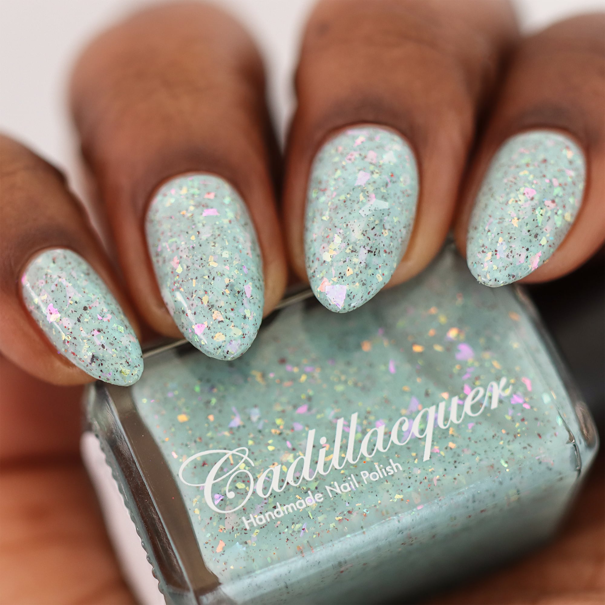Cadillacquer Fresh Sheets pastel mint iridescent crelly nail polish, bottle held with four-nail swatch, deep skin, studio lighting