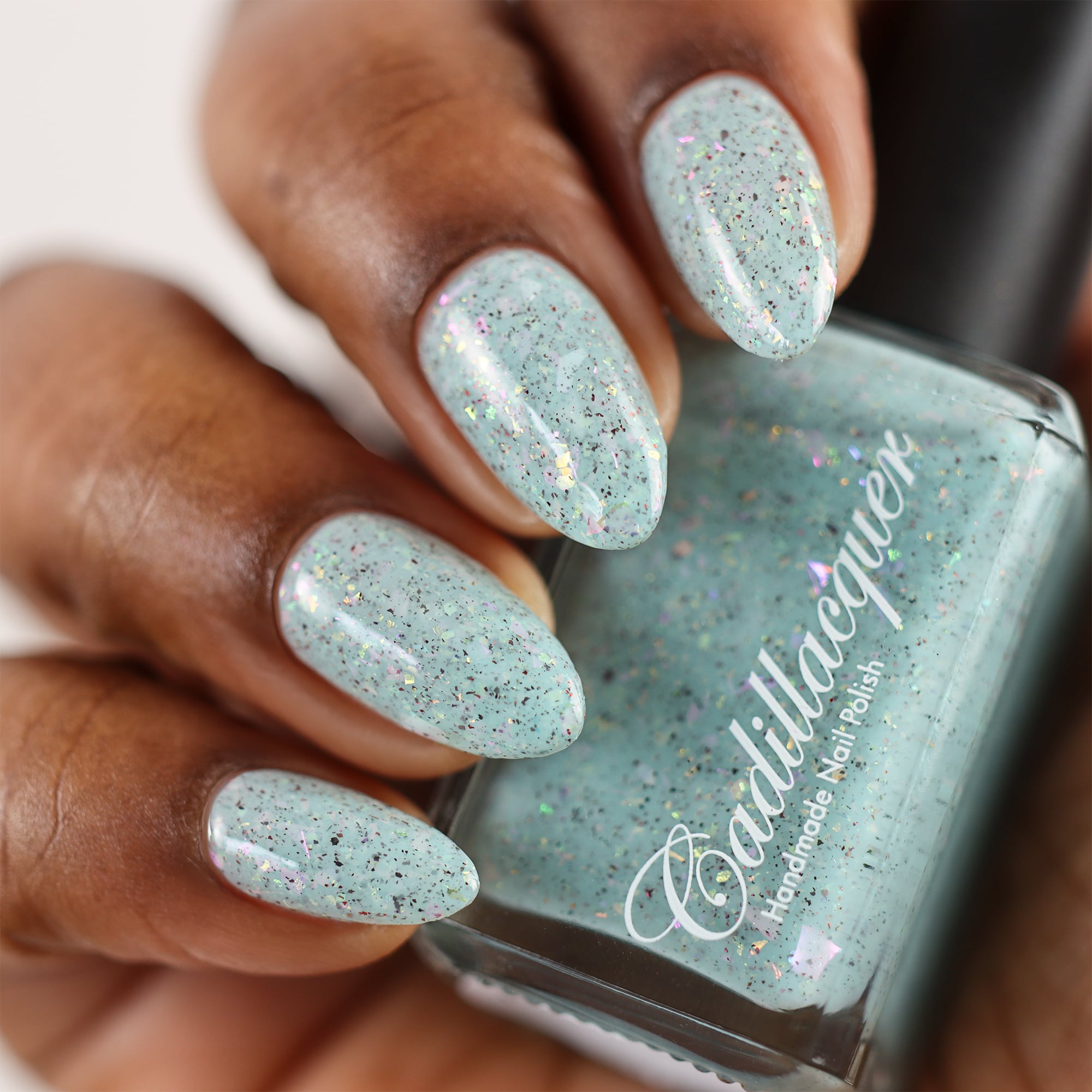 Cadillacquer Fresh Sheets pastel mint iridescent crelly nail polish, bottle held with four-nail swatch, rich skin, studio lighting