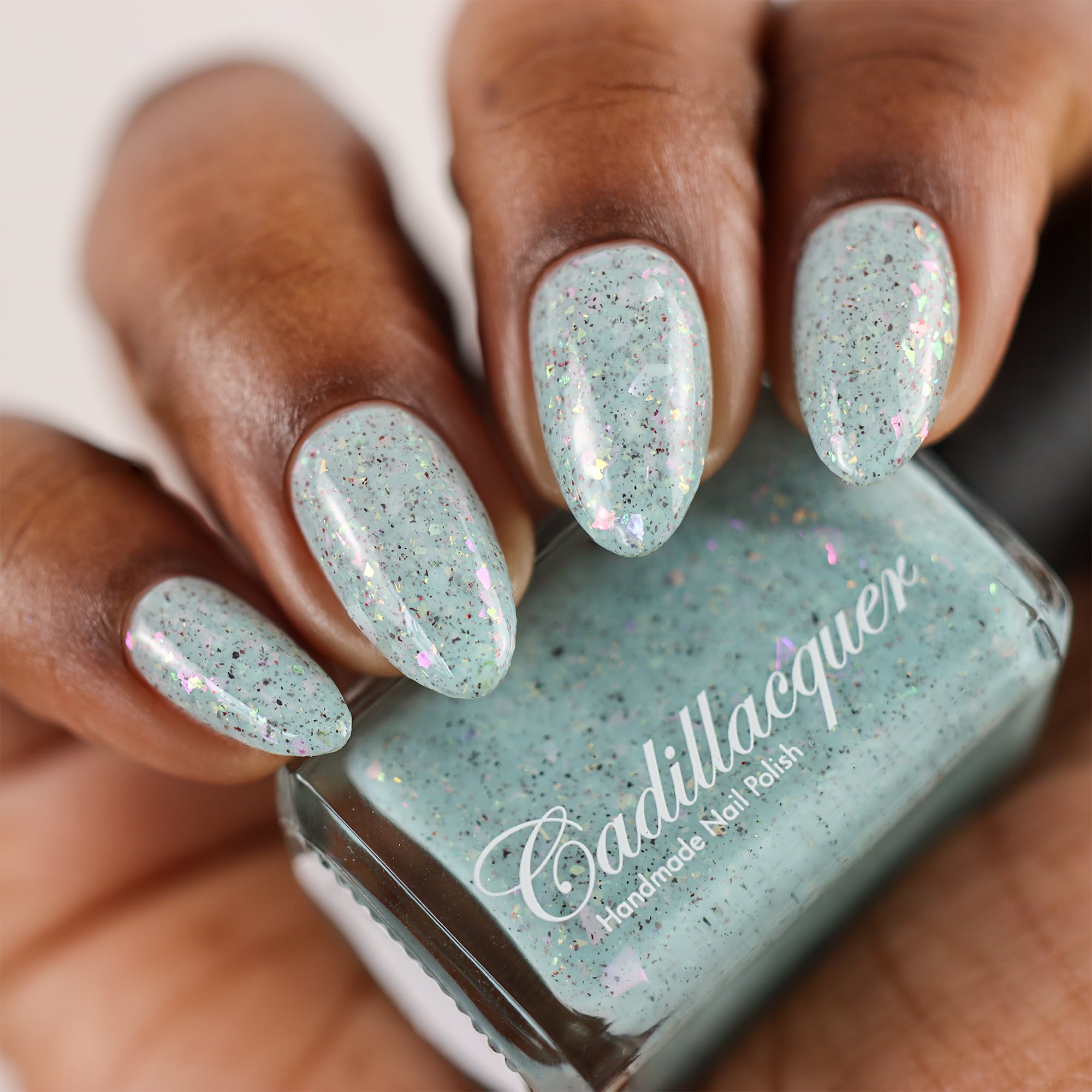 Cadillacquer Fresh Sheets pastel mint iridescent crelly nail polish, bottle held with four-nail swatch, deep skin, studio lighting
