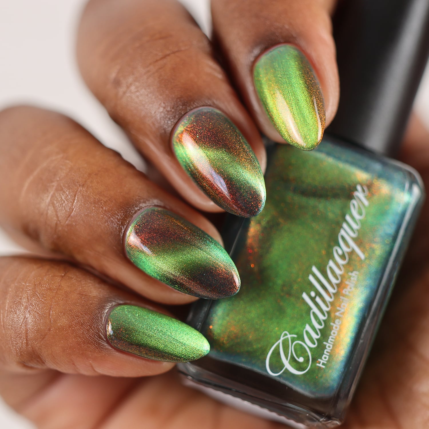 Magnetic multichrome aurora shimmer Cadillacquer nail polish in I Found A Place, five almond-shaped nails on rich skin tone showing a shifting lime-green magnetic band over a dark olive-teal field base with vivid red-to-orange aurora shimmer flakes throughout, held alongside the teal-green bottle, studio lighting, swatched by @doseoflolade