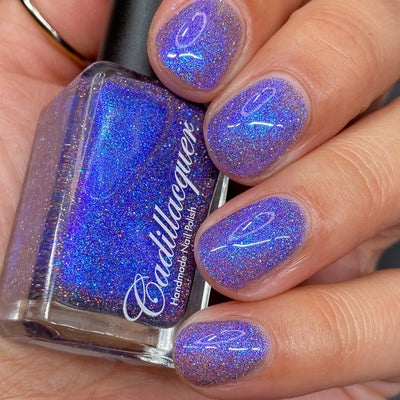 Holographic shimmer and micro-flake Cadillacquer nail polish in I'll Be Right Back, shown as bottle held in hand with five swatched nails, periwinkle-violet base densely packed with electric blue shimmer, blue micro flakes, red and charcoal holographic glitter sparks, light-medium skin tone, studio lighting, swatched by @heeymercedes