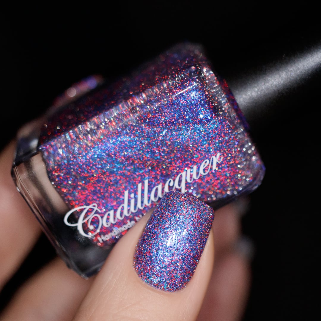 Shimmer, glitter and micro-flake Cadillacquer nail polish in I'll Be Right Back, shown as a bottle-in-hand with one swatched nail, the polish is a deep blue-violet base loaded with red and charcoal reflective glitter, blue micro flakes and blue shimmer flash, light skin tone, studio lighting against black background, swatched by @yyulia_m