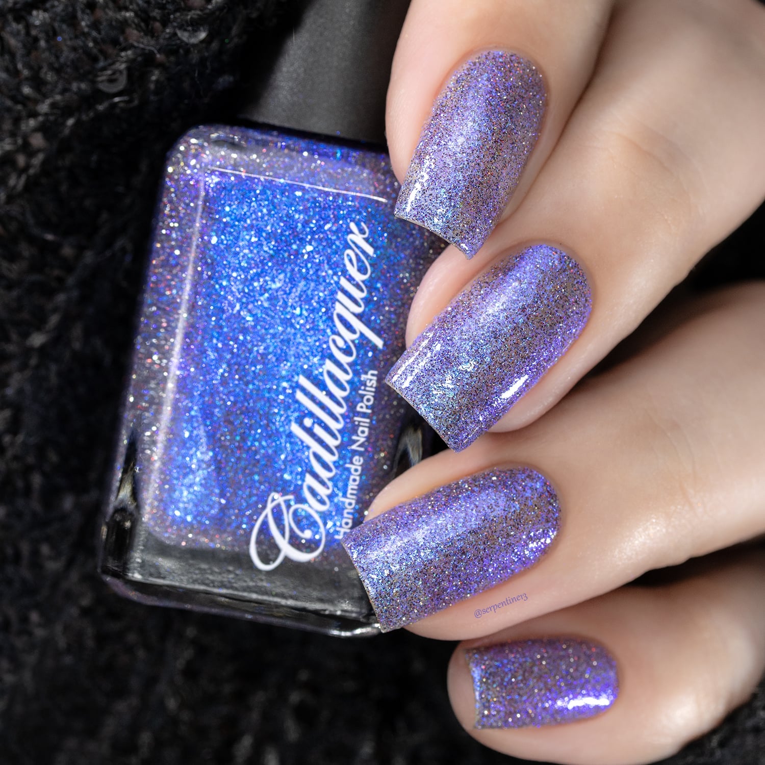 Holographic shimmer Cadillacquer nail polish in I'll Be Right Back, shown in bottle held in hand with four square nails swatched, polish appears lavender-violet on nails with dense blue micro flake shimmer and scattered holographic glitter sparks in charcoal and red, bottle face reads bright cerulean blue under studio lighting, light skin tone, swatched by @serpentine13