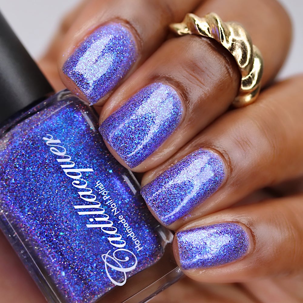 Holographic shimmer and micro-flake Cadillacquer nail polish in I'll Be Right Back, shown in bottle held against four swatched nails on deep skin tone, blue-violet base packed with pale periwinkle shimmer flash, iridescent blue micro flakes, and red and charcoal reflective glitter sparks, under studio lighting, swatched by @lacquer_is_life