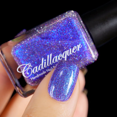 Holographic shimmer and micro-flake Cadillacquer nail polish in I'll Be Right Back, shown as a bottle held in hand with one swatched nail visible, violet-purple base packed with electric blue shimmer, fine blue micro flakes and holographic scatter, light-medium skin tone, studio lighting, swatched by @yyulia_m