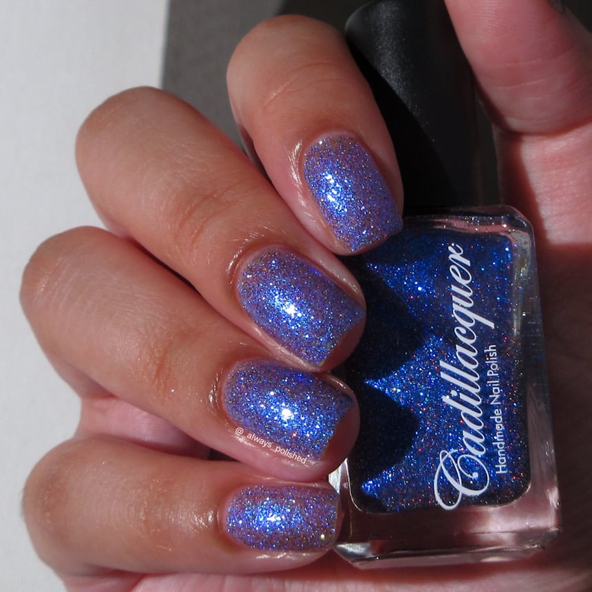 Shimmer, micro-flake and reflective glitter Cadillacquer nail polish in I'll Be Right Back, shown on four short squoval nails on a light-medium skin tone hand holding the deep royal blue bottle, in bright daylight lighting, swatched by @_always_polished_