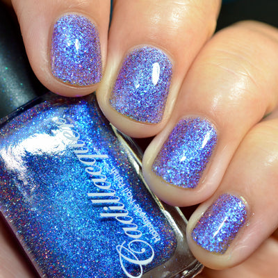 Holographic glitter and blue shimmer flake Cadillacquer nail polish in I'll Be Right Back, shown on five short natural nails on a light-medium skin tone hand holding the bottle, bottle appears vivid electric blue with holographic flash while nails read as sheer lavender-periwinkle densely packed with blue micro flakes and scattered red-pink and charcoal holographic glitter sparks, direct flash indoor lighting, swatched by @caitlinswatches