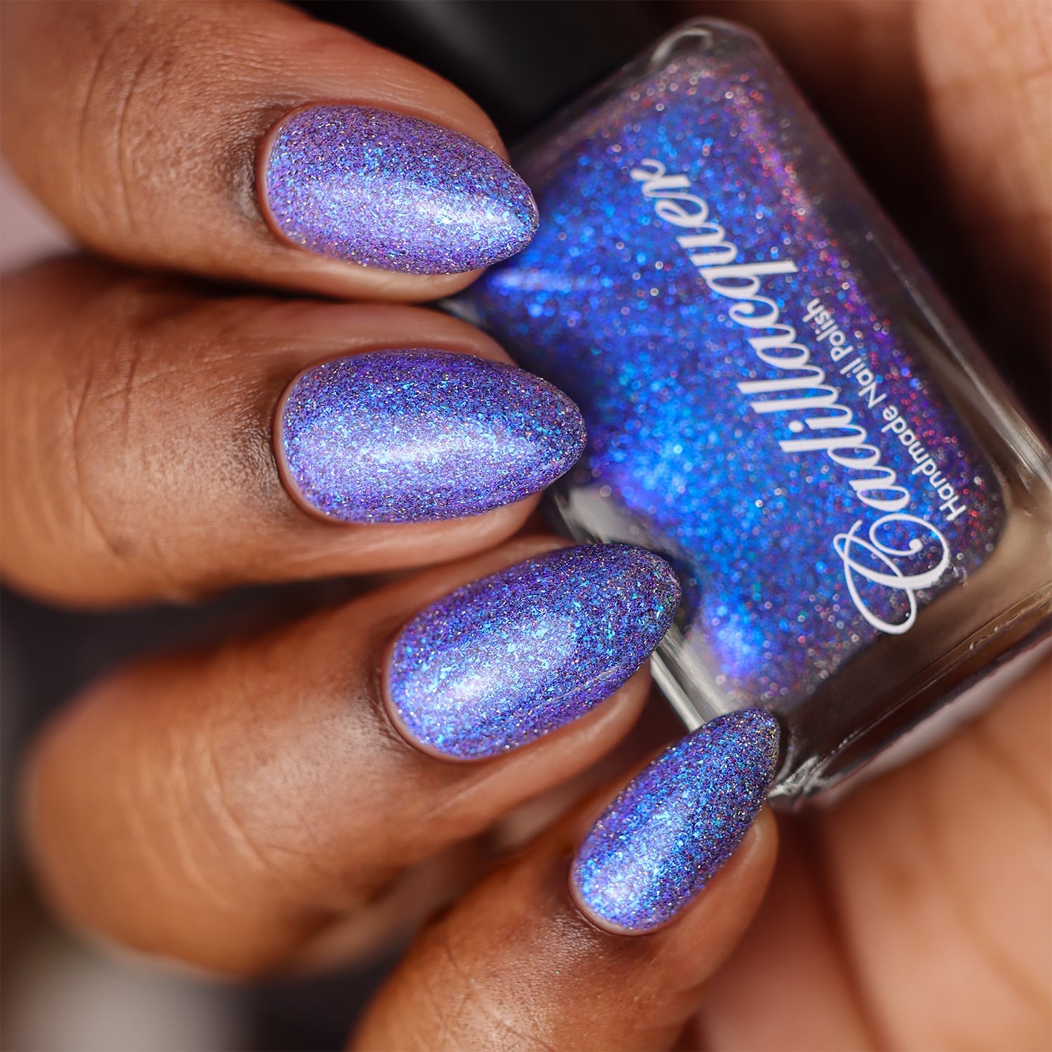 Holographic shimmer and micro-flake Cadillacquer nail polish in I'll Be Right Back, shown as a swatch on five almond-shaped nails and held bottle, shifting from periwinkle violet to cornflower blue with dense blue micro flakes and scattered red and charcoal reflective glitter particles, on deep skin tone, studio lighting, swatched by @doseoflolade