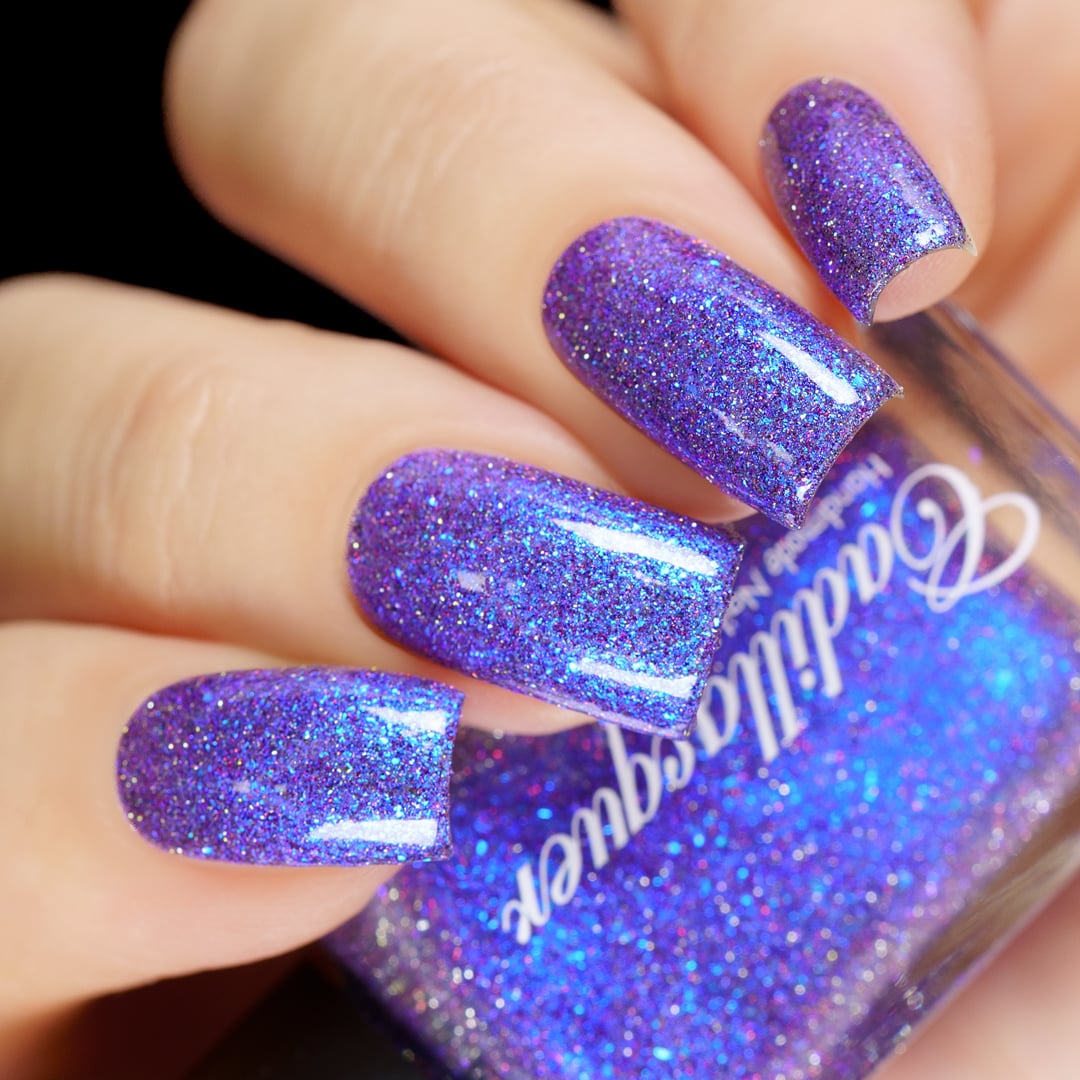 Holographic shimmer and micro-flake Cadillacquer nail polish in I'll Be Right Back, shown as a swatch on four almond-shaped nails held alongside the bottle, displaying a vivid violet-purple base densely packed with blue micro flakes, blue shimmer, and charcoal and red holographic glitter particles that scatter rainbow light, on light skin tone, under direct flash lighting, swatched by @yyulia_m