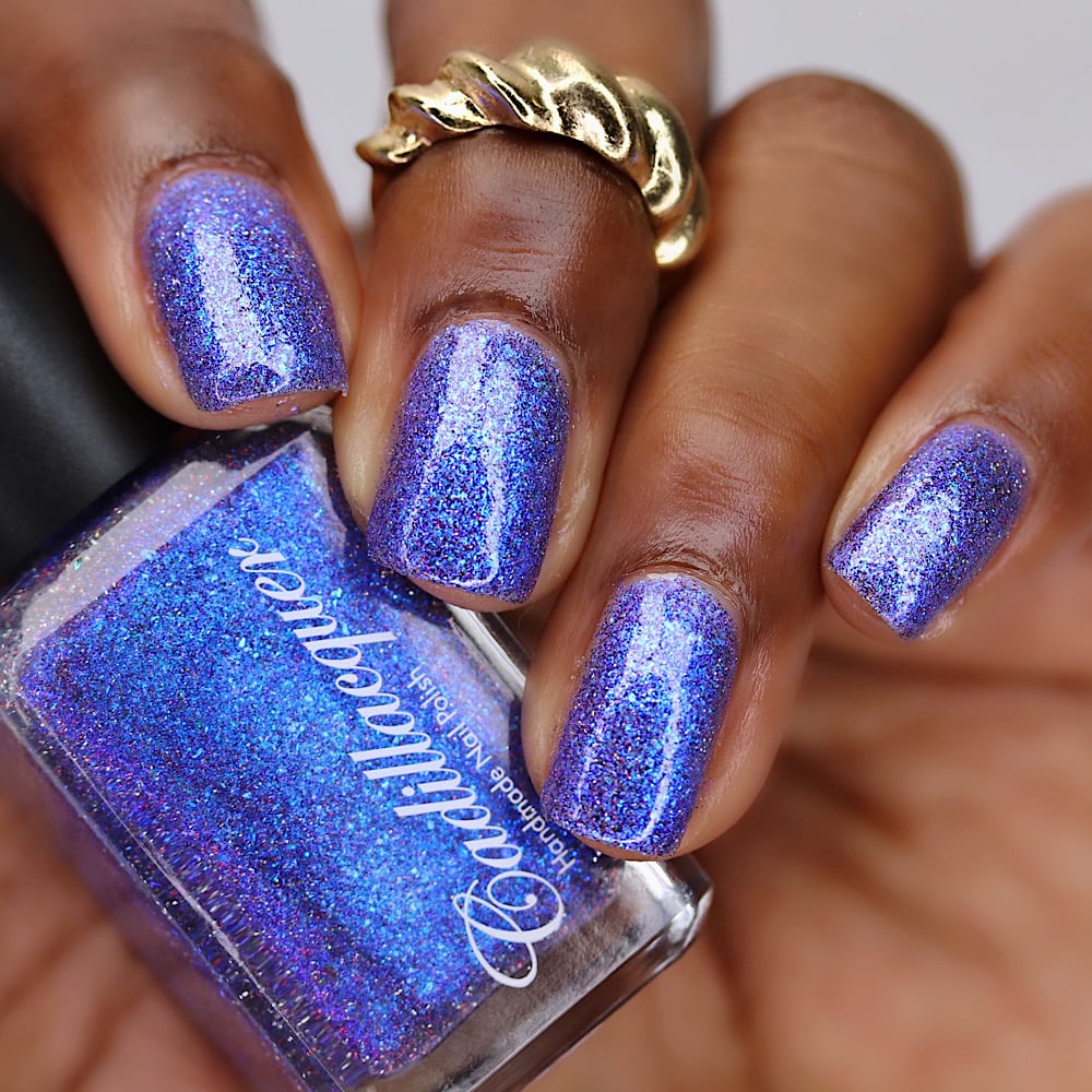 Holographic shimmer and micro-flake Cadillacquer nail polish in I'll Be Right Back, shown swatched on eight short square nails held around the bottle, with deep skin tone, studio lighting, the polish reads as blue-violet with electric blue shimmer flash and fine iridescent flakes packed throughout, swatched by @lacquer_is_life