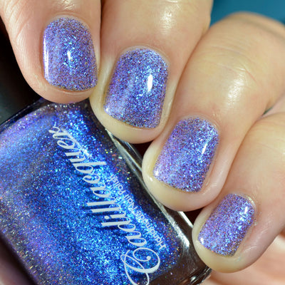 Shimmer, micro flake and holographic glitter Cadillacquer nail polish in I'll Be Right Back, shown as a bottle-and-nails swatch with five square nails reading sheer lavender-violet packed with blue iridescent micro flakes and sparkling holographic glitter, while the bottle reads vivid electric blue, on light skin tone under studio lighting, swatched by @caitlinswatches