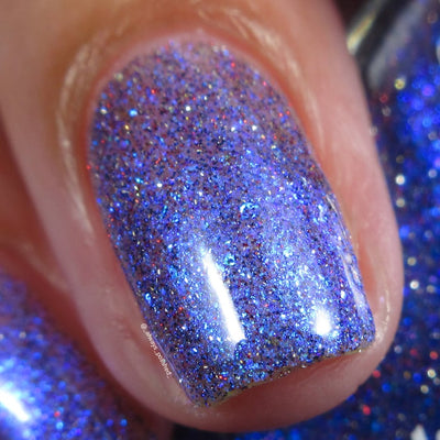 Shimmer, fine flake and reflective glitter Cadillacquer nail polish in I'll Be Right Back, shown as a macro close-up swatch on two short natural nails with light skin tone, displaying a sheer periwinkle-violet base densely packed with blue micro flakes, bright electric-blue shimmer flash, and scattered red and charcoal holographic glitter sparks, under direct flash lighting, swatched by @_always_polished_