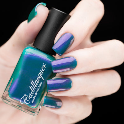 Multichrome shimmer Cadillacquer nail polish in It's All For You, shown in bottle held in hand with nails swatched, shifting from deep teal green at edges to vivid violet-purple across the nail face with lavender shimmer flash, light skin tone with squoval nails, studio lighting on black background, swatched by @rafinails