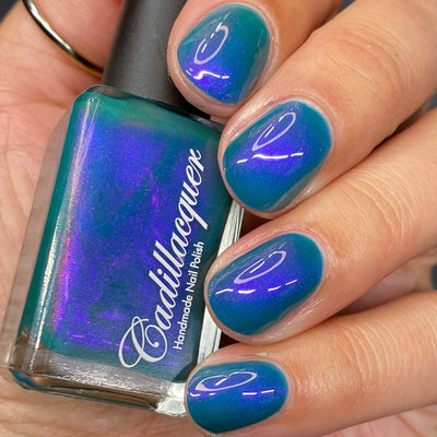 Multichrome shimmer Cadillacquer nail polish in It's All For You, shown as a bottle held against five swatched nails shifting from teal green to violet-purple depending on angle, light-medium skin tone, bright daylight lighting, swatched by @heeymercedes
