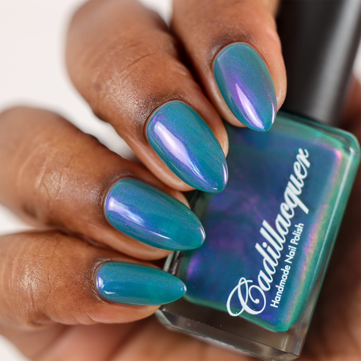 Shimmer multichrome Cadillacquer nail polish in It's All For You, shown on four almond-shaped nails on deep skin tone holding the bottle, displaying a teal green base packed with shifting lavender to violet purple shimmer, studio lighting, swatched by @doseoflolade