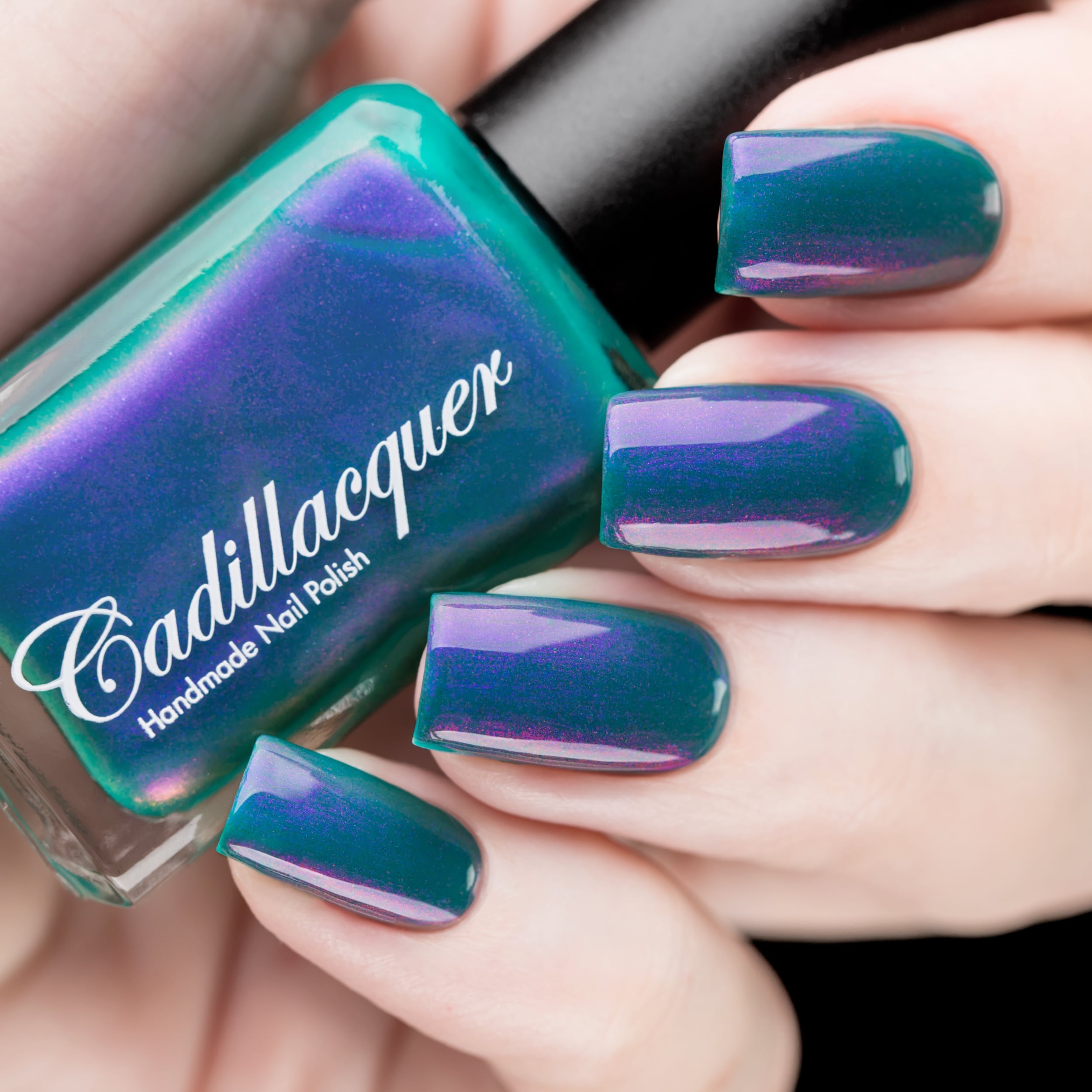 Multichrome shimmer Cadillacquer nail polish in It's All For You, shown as a bottle held in hand beside five swatched squoval nails, shifting from deep teal green base to violet-purple and soft lavender at different angles, on light skin tone, under studio lighting, swatched by @rafinails