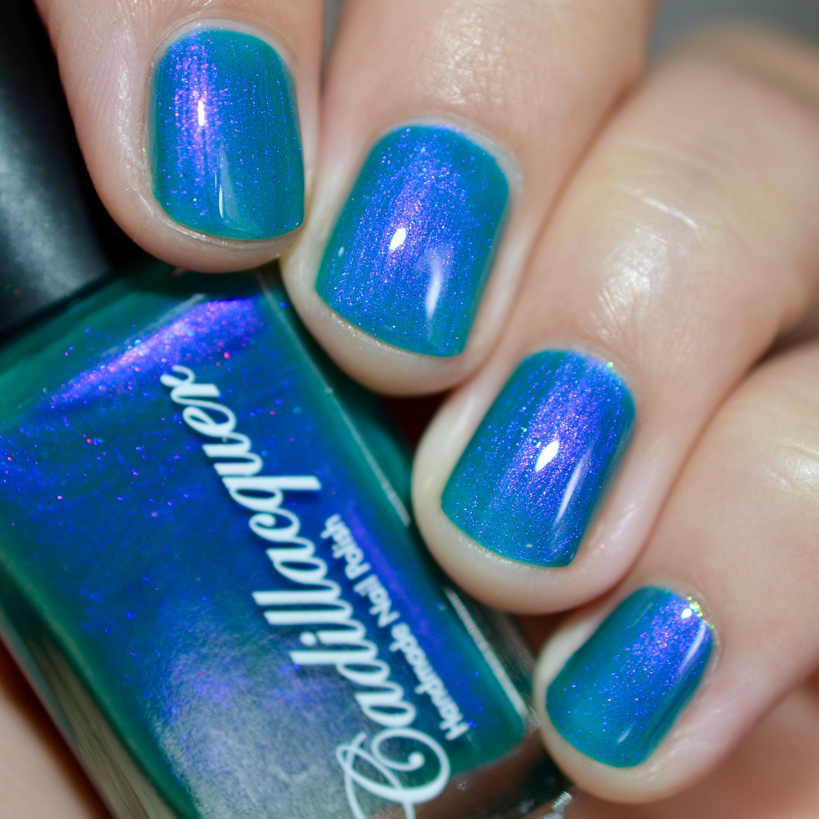 Shimmer multichrome Cadillacquer nail polish in It's All For You, shown swatched on four short squoval nails alongside the bottle, teal-green base shifts through blue-violet to pink-purple shimmer at different angles, light-medium skin tone, studio lighting, swatched by @caitlinswatches