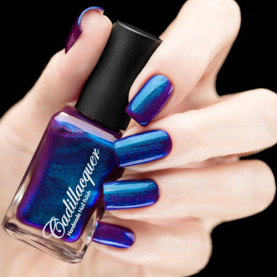 Multichrome shimmer Cadillacquer nail polish in Keep Doubting, shown as a bottle held in hand with five swatched nails visible, the bottle reads deep mulberry-violet with intense blue-teal shimmer across the face, while the nails shift from deep indigo-blue to teal and purple depending on angle, light skin tone, studio lighting, swatched by @rafinails