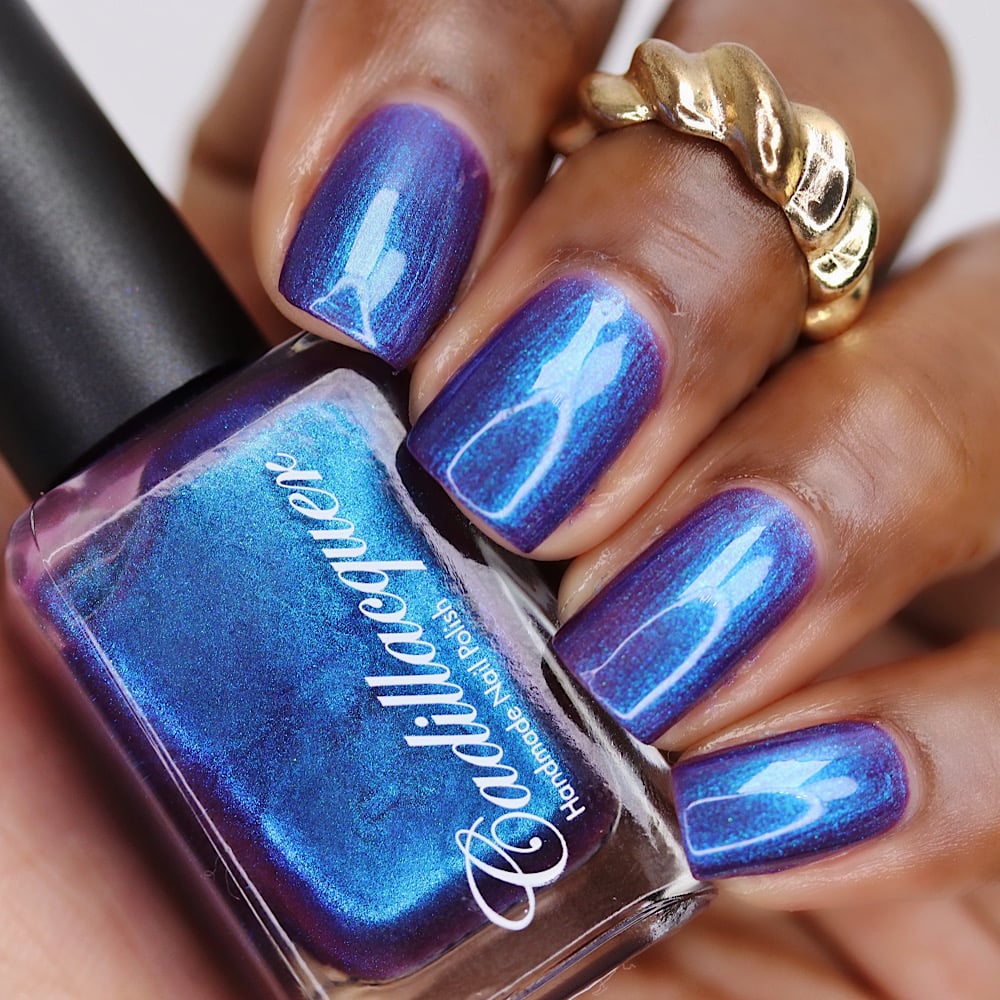 Multichrome shimmer Cadillacquer nail polish in Keep Doubting, shown in bottle held in hand alongside four swatched nails, the deep violet-indigo base shifts to vivid cerulean and icy blue shimmer flash at different angles, deep skin tone with gold twisted ring accent, studio lighting, swatched by @lacquer_is_life