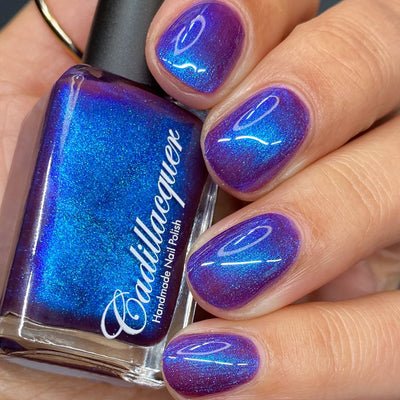 Shimmer Cadillacquer nail polish in Keep Doubting, shown as a square bottle held in hand alongside five swatched nails, the bottle face blazes electric blue-teal with shimmer while the nails reveal a deep mulberry-violet base with vivid cobalt blue shimmer flash shifting to teal-green at steep angles, light-medium skin tone, studio lighting, swatched by @heeymercedes