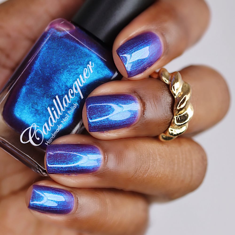 Shimmer multichrome Cadillacquer nail polish in Keep Doubting, shown in bottle held in hand with four swatched square nails shifting from deep blue-violet to electric cobalt blue, worn on deep skin tone with a gold twisted ring, studio lighting, swatched by @lacquer_is_life