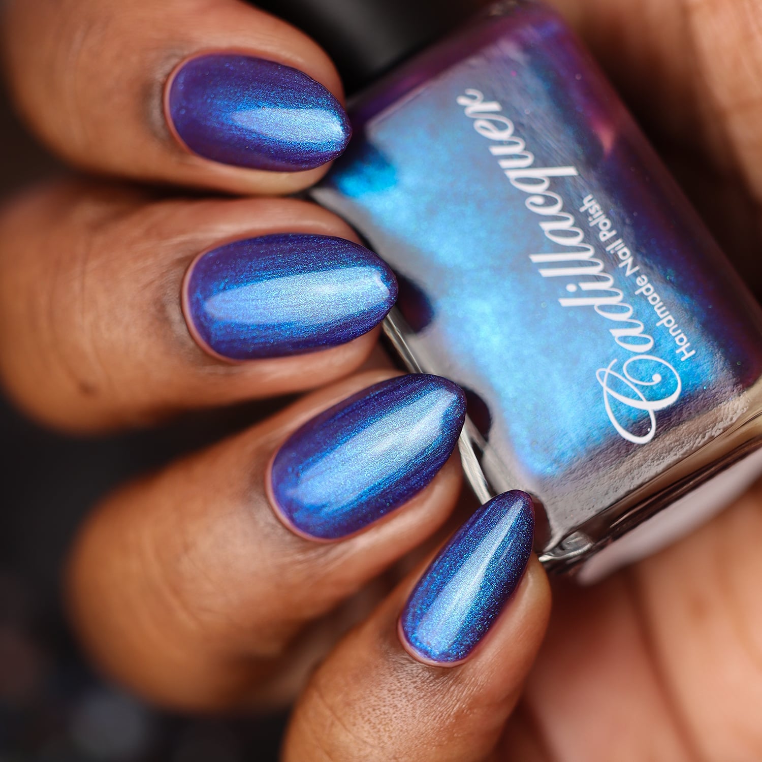Multichrome shimmer Cadillacquer nail polish in Keep Doubting, shown in a bottle-in-hand swatch with five almond-shaped nails on deep skin tone, the polish shifts from deep blue-violet in shadow to bright periwinkle and electric cyan-blue under direct light, with the glass bottle face dominated by vivid cyan shimmer, studio lighting, swatched by @doseoflolade