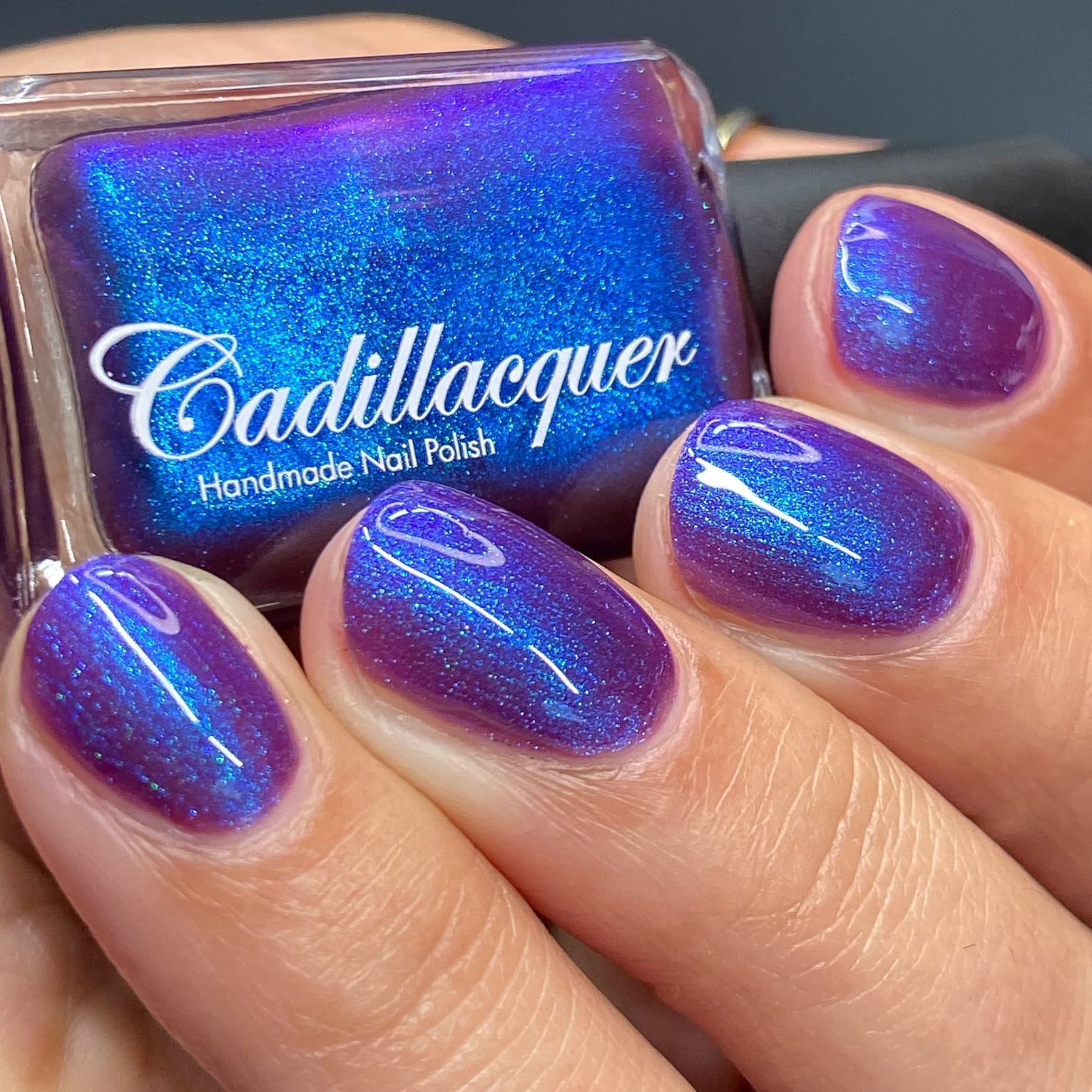 Shimmer Cadillacquer nail polish in Keep Doubting, shown on four short oval nails with a matching bottle held in hand, deep violet-mulberry base packed with vivid electric blue and teal shimmer that flashes intensely at angles, light-medium skin tone, studio lighting, swatched by @heeymercedes