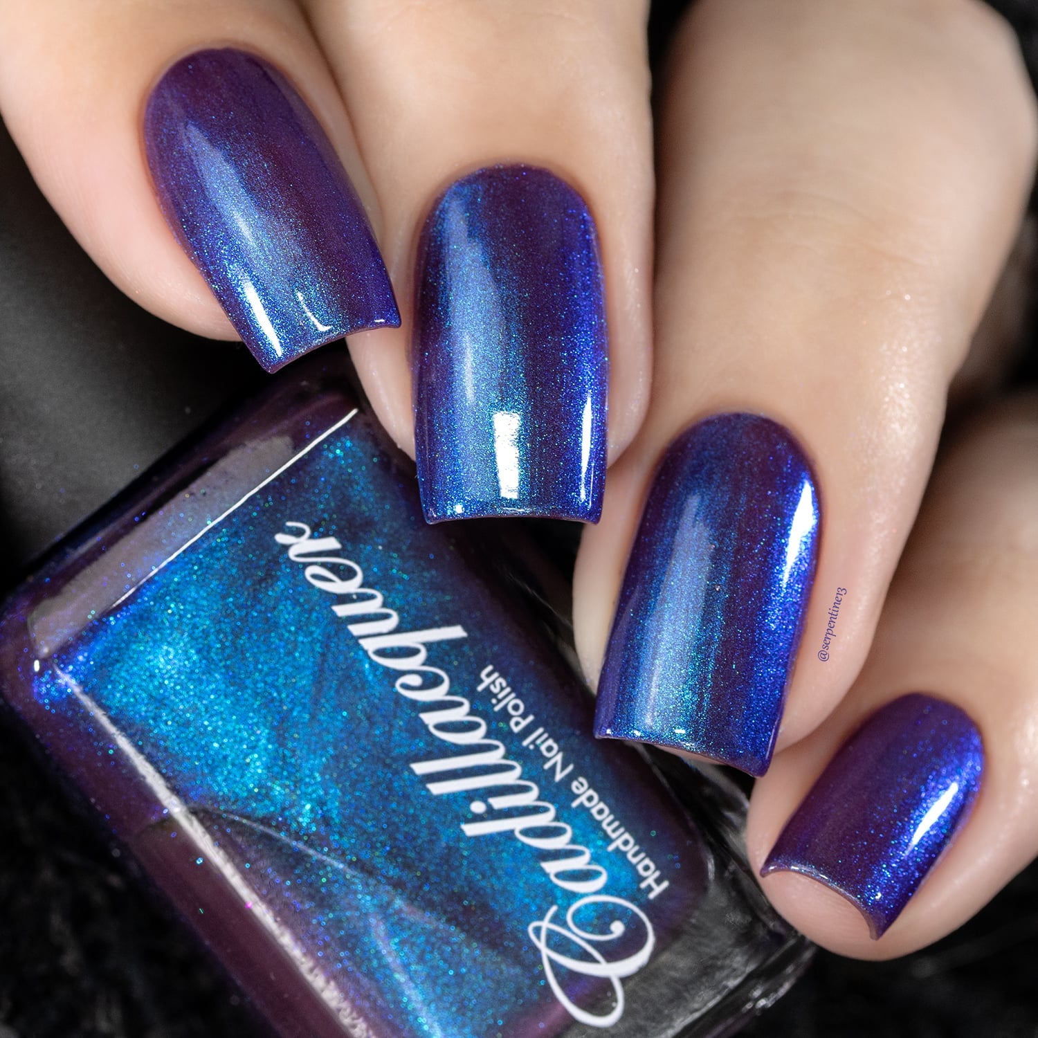 Holographic multichrome shimmer Cadillacquer nail polish in Keep Doubting, shown as a swatch on four squoval nails with a bottle propped against the hand, the deep mulberry-violet base shifts through cobalt blue and cyan-teal shimmer depending on angle, light skin tone, studio lighting, swatched by @serpentine13