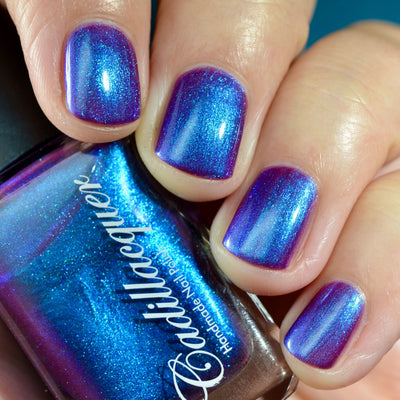 Multichrome shimmer Cadillacquer nail polish in Keep Doubting, shown swatched on four short natural nails held around a Cadillacquer bottle, nails shift from deep violet-purple at face-on angles to electric blue and mulberry-magenta at oblique angles with vivid blue shimmer throughout, light skin tone, studio lighting, swatched by @caitlinswatches