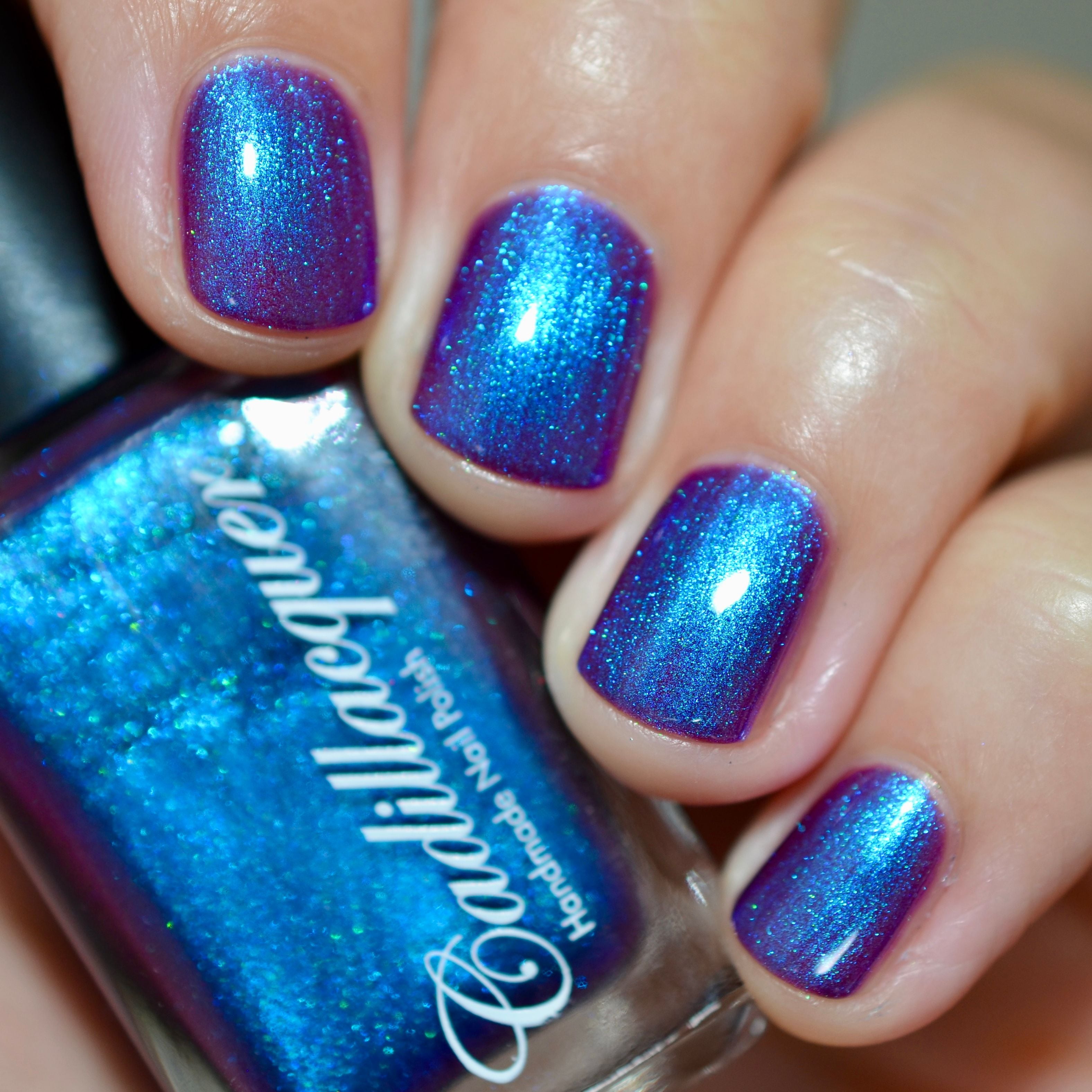 Multichrome shimmer Cadillacquer nail polish in Keep Doubting, shown on four short natural nails and held alongside a Cadillacquer bottle, the polish shifts from deep violet-purple at face-on angles through vivid cerulean blue shimmer to warm magenta-pink at raking angles, with a teal-green micro shimmer visible in macro, light skin tone, direct flash lighting, swatched by @caitlinswatches