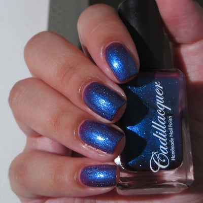 Shimmer Cadillacquer nail polish in Keep Doubting, swatched on four short natural nails held alongside the polish bottle, showing a deep mulberry-purple base densely packed with vivid cobalt-blue shimmer that flares brilliantly at angular highlights, on light skin tone, studio lighting, swatched by @_always_polished_