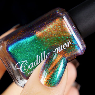 Holographic magnetic multichrome Cadillacquer nail polish in Once You See It, shown in bottle held in hand with single square nail swatch displaying teal-green magnetic band over deep copper-rust field with aurora green-to-blue shimmer scatter, light skin tone, studio lighting, swatched by @yyulia_m