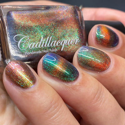 Magnetic holographic multichrome Cadillacquer nail polish in Once You See It, shown in bottle held in hand with five swatched nails displaying a deep red-brown base with a vivid amber-orange magnetic cat-eye band and green-to-blue aurora shimmer shifts, light skin tone, studio lighting, swatched by @heeymercedes