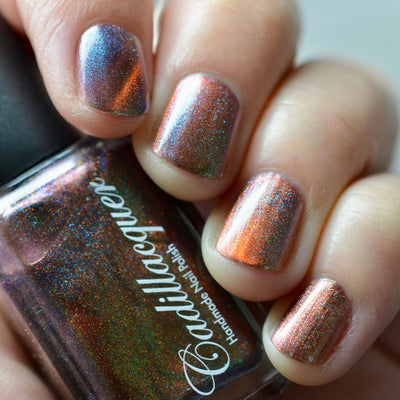 Holographic magnetic multichrome Cadillacquer nail polish in Once You See It, shown in bottle held in hand alongside four short square nails swatched with a copper-orange magnetic band over a dark reddish-brown field with green-to-blue aurora shimmer, light skin tone, bright daylight lighting, swatched by @caitlinswatches