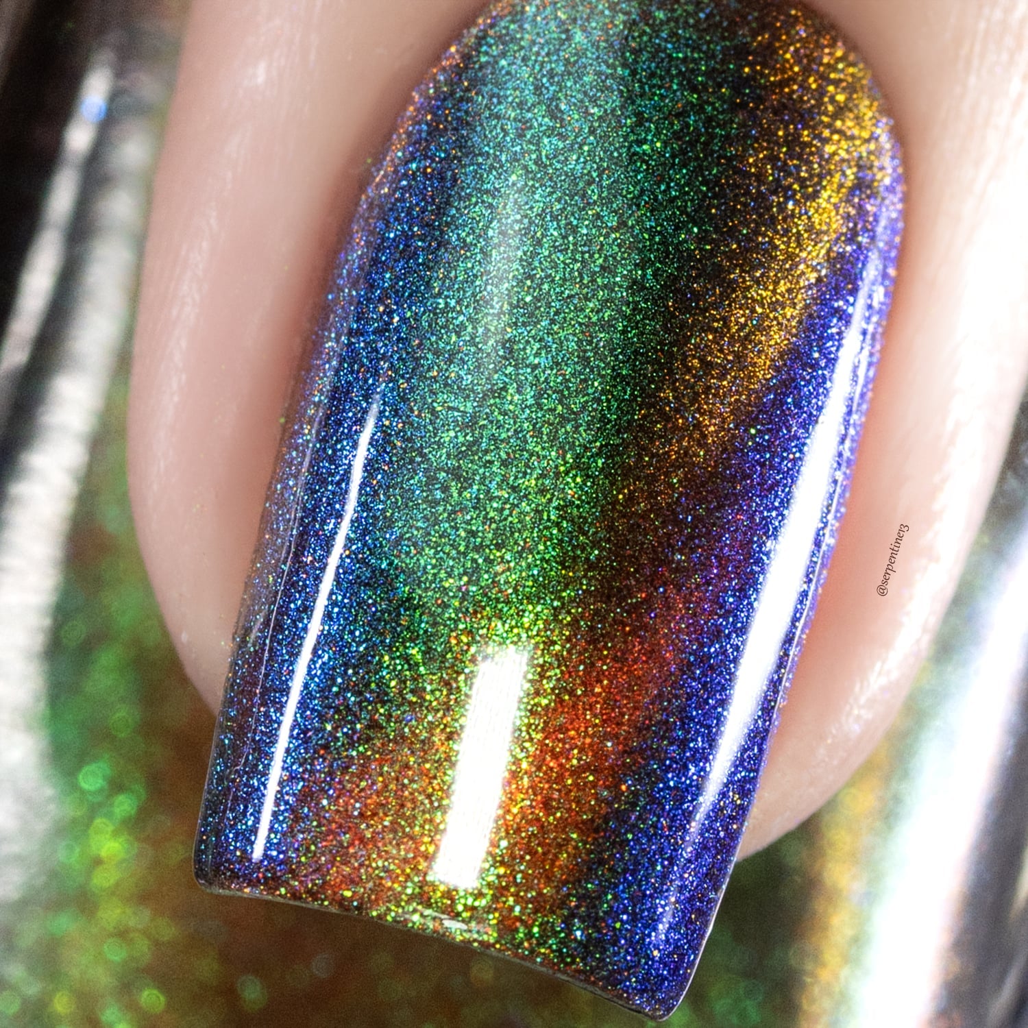 Holographic magnetic multichrome Cadillacquer nail polish in Once You See It, extreme macro closeup of a single square nail showing a deep navy-indigo carrier base scattered with ultra-fine holographic particles that flash green, teal, amber-gold, burnt orange, and violet across the nail surface, with a diagonal amber-gold magnetic band concentrated across the centre, on light skin tone, studio lighting, swatched by @serpentine13