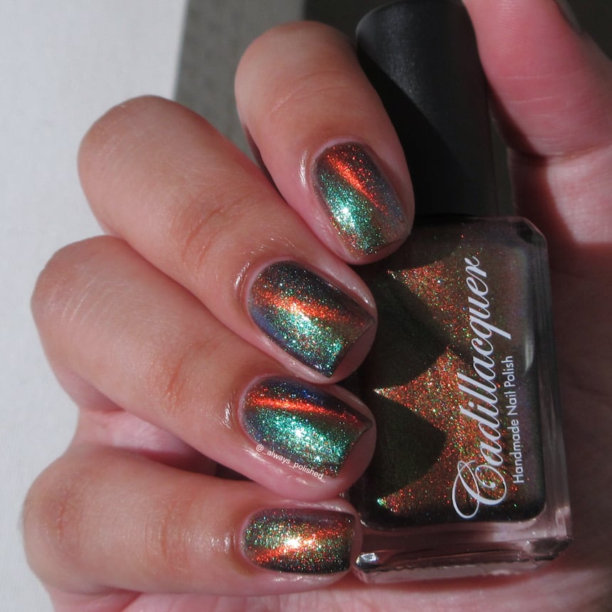 Magnetic multichrome Cadillacquer nail polish in Once You See It, swatched on four short natural nails held alongside the bottle, showing a deep copper-orange diagonal magnetic band over a very dark olive-black field with vivid teal-green to blue aurora shimmer throughout, on light skin tone, in bright daylight, swatched by @_always_polished_