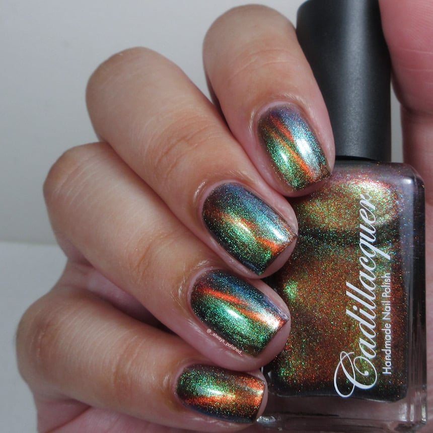 Magnetic multichrome shimmer Cadillacquer nail polish in Once You See It, shown on eight short natural nails held alongside the bottle, deep forest-green field with vivid burnt-orange magnetic stripe and green-to-teal aurora shimmer flash, light skin tone, studio lighting, swatched by @_always_polished_