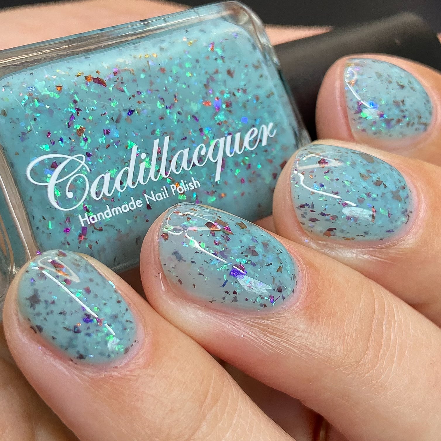 Cadillacquer Pastel Thoughts pastel teal purple chameleon iridescent crelly nail polish, bottle held with four-nail swatch, light skin, studio lighting