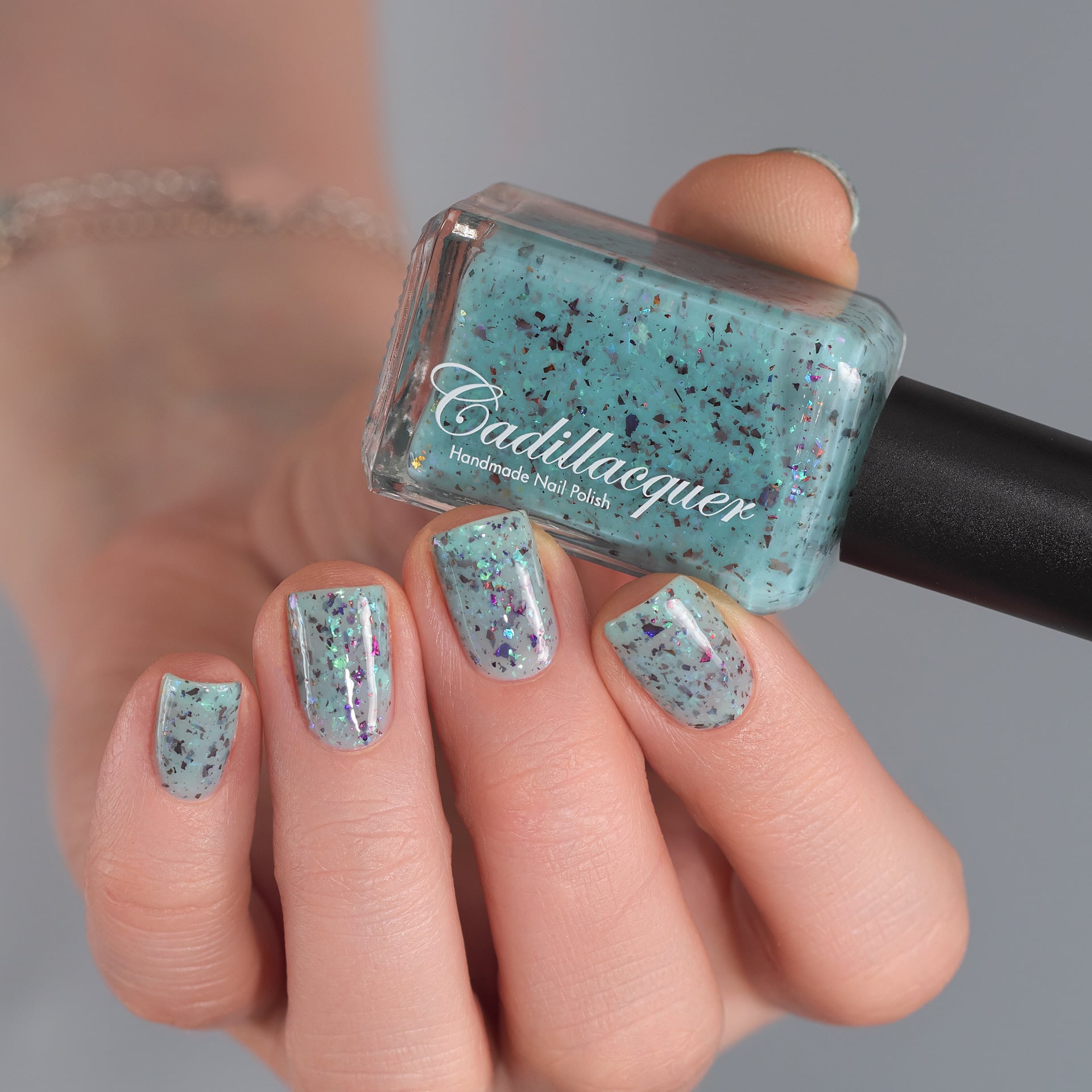 Cadillacquer Pastel Thoughts pastel teal purple chameleon iridescent crelly nail polish, bottle held with four-nail swatch, light skin, studio lighting