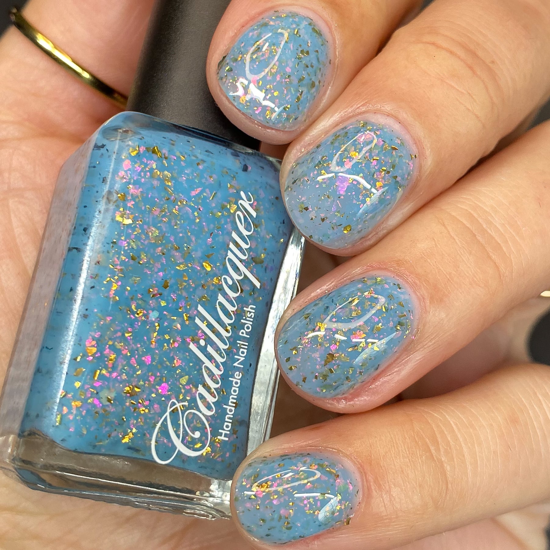 Cadillacquer Slow Mornings light blue gold chameleon iridescent crelly nail polish, bottle held with four-nail swatch, light skin, bright daylight lighting